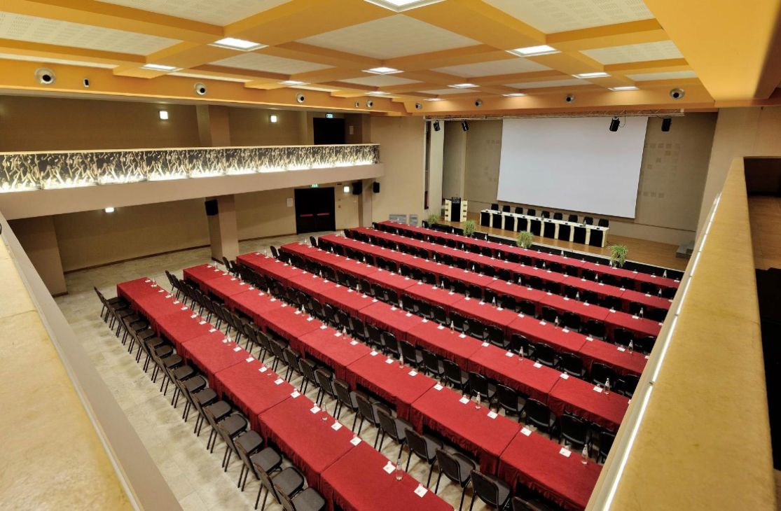 Sala conferenze con tavoli e sedie disposti ordinatamente, grande schermo e podio.