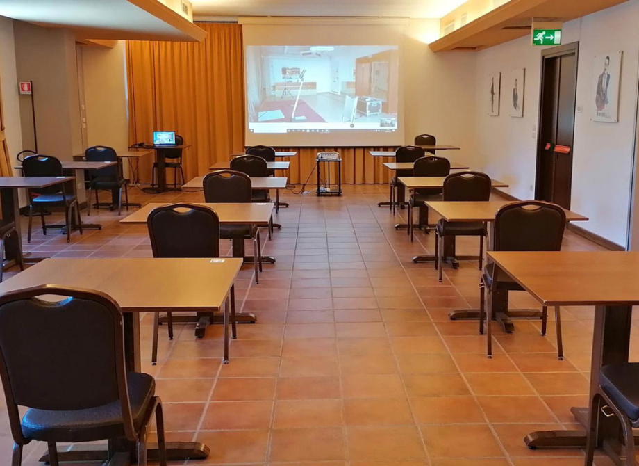 Aula conferenze con tavoli distanziati e proiettore acceso.