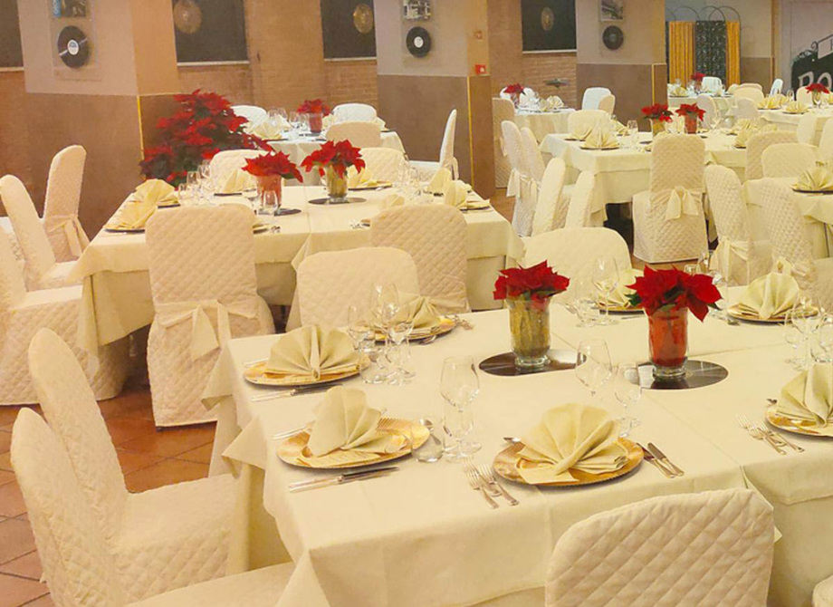 Sala elegante con tavoli apparecchiati, sedie coperte e centrotavola di fiori rossi.