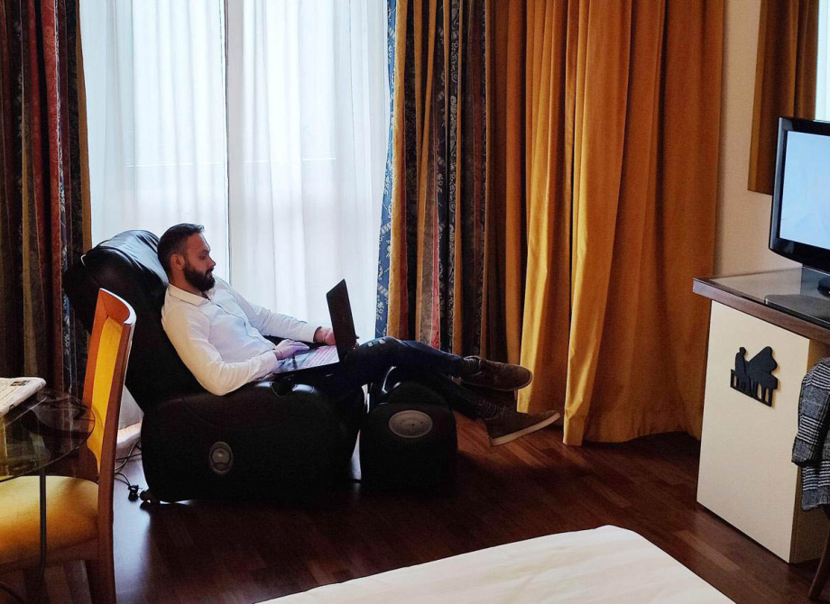 Uomo su poltrona massaggiante con laptop, TV accesa, stanza con tende e lampada jazz.