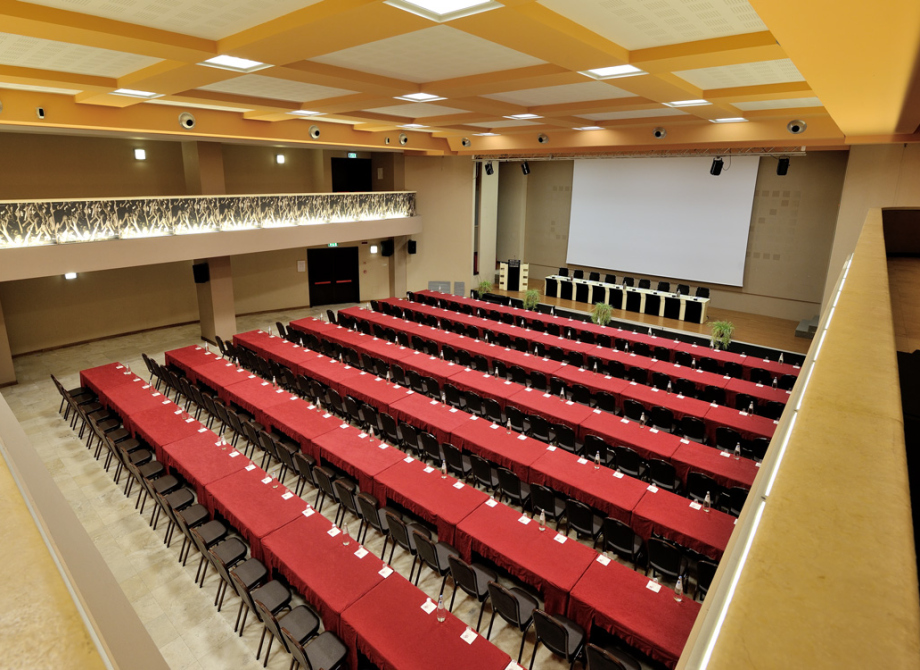 Ampia sala conferenze con file di tavoli rossi e grande schermo sullo sfondo.