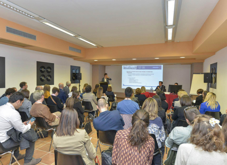 Conferenza con pubblico attento, presentazione su schermo, ambiente formale e professionale.