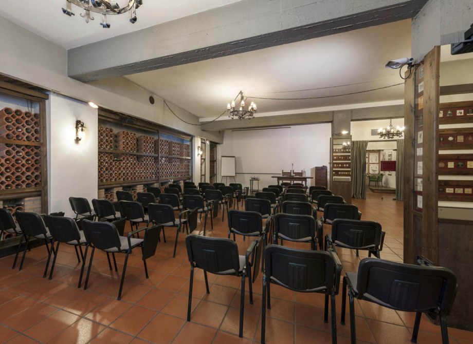Sala conferenze con sedie nere, scaffali di bottiglie e illuminazione a lampadario.