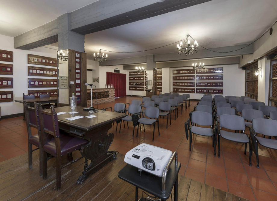 Sala conferenze con sedie, proiettore e tavolo con microfono, illuminata da lampadari.