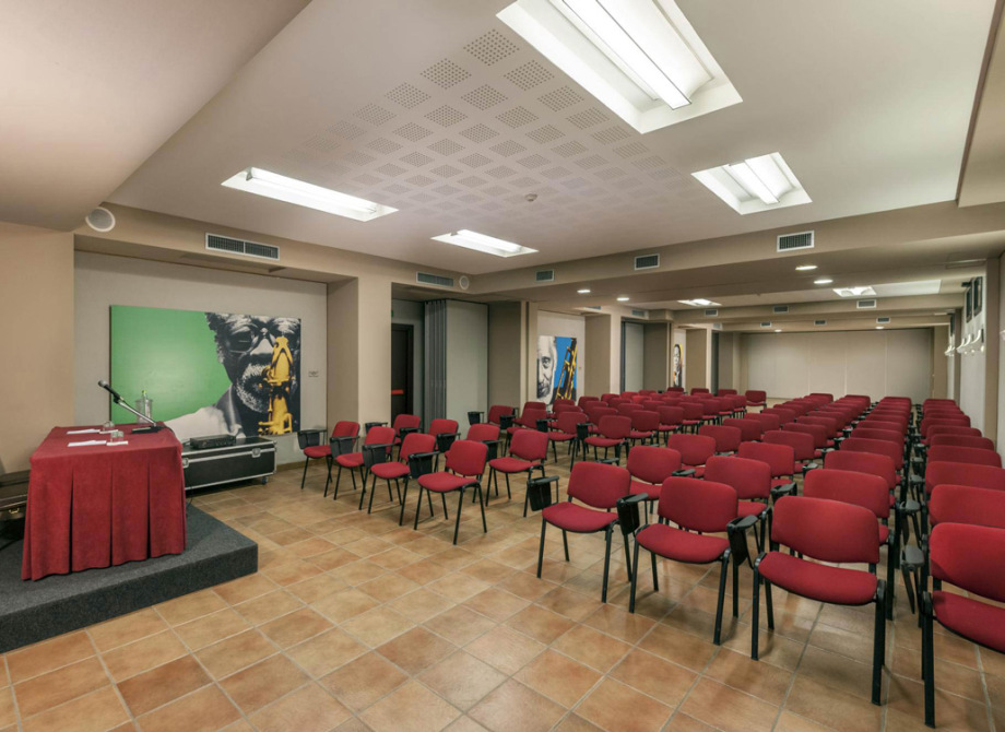 Sala conferenze con sedie rosse, podio e opere d'arte alle pareti.